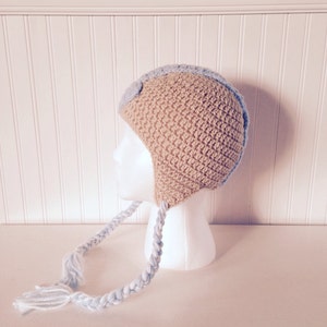 May include: A crocheted baby hat with a blue and beige color scheme. The hat has a long braided strap with a blue and white color scheme.