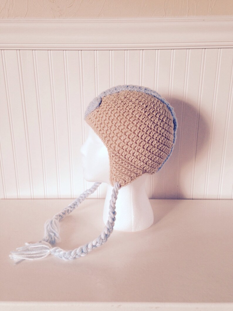 May include: A crocheted baby hat with a blue and beige color scheme. The hat has a long braided strap with a blue and white color scheme.