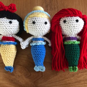 Pu&ograve; includere: Tre bambole sirene all'uncinetto con capelli e code di diversi colori. La bambola a sinistra ha i capelli neri e una coda gialla, la bambola al centro ha i capelli biondi e una coda blu, e la bambola a destra ha i capelli rossi e una coda verde.