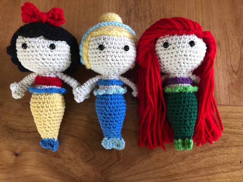 Pu&ograve; includere: Tre bambole sirene all'uncinetto con capelli e code di diversi colori. La bambola a sinistra ha i capelli neri e una coda gialla, la bambola al centro ha i capelli biondi e una coda blu, e la bambola a destra ha i capelli rossi e una coda verde.