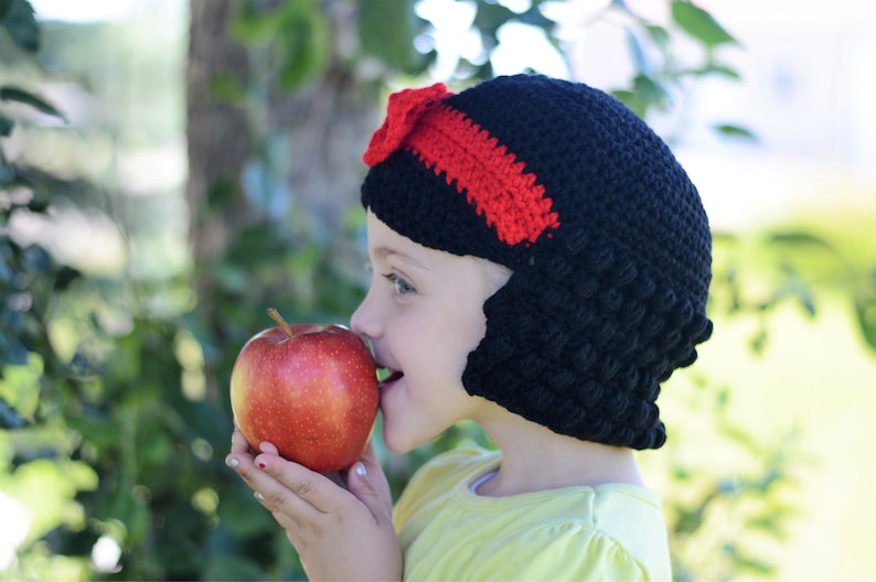 Puede incluir: Un gorro negro de ganchillo con un lazo y una diadema rojos, que recuerda el peinado de un personaje. Una persona sostiene una manzana roja. El dise&ntilde;o del gorro evoca a un personaje de cuento de hadas.