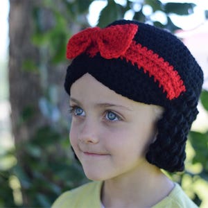 Puede incluir: Un gorro negro de ganchillo con un lazo y una raya rojos, que se asemeja al peinado de un personaje. El gorro tiene pelo negro de ganchillo en los lados. El gorro lo lleva una persona con ojos azules.