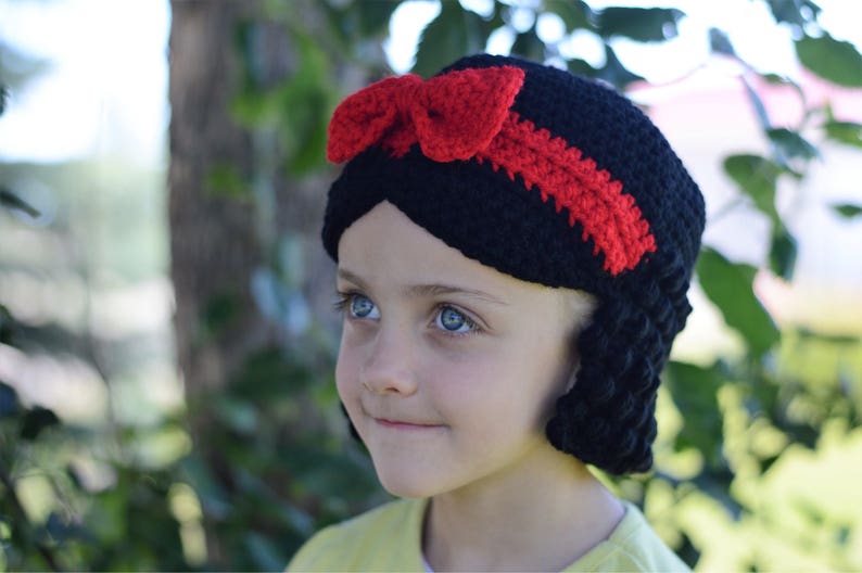 Puede incluir: Un gorro negro de ganchillo con un lazo y una raya rojos, que se asemeja al peinado de un personaje. El gorro tiene pelo negro de ganchillo en los lados. El gorro lo lleva una persona con ojos azules.
