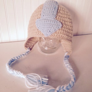 May include: A crocheted baby hat with a blue and white braid and tassels. The hat features a blue and white crocheted design on the top, resembling the Avatar symbol.