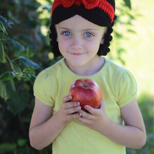 Puede incluir: Una diadema negra de ganchillo con lazo y ribete rojos, combinada con una camiseta amarilla y una manzana roja. La diadema tiene un dise&ntilde;o &uacute;nico, y la manzana se sostiene en las manos.
