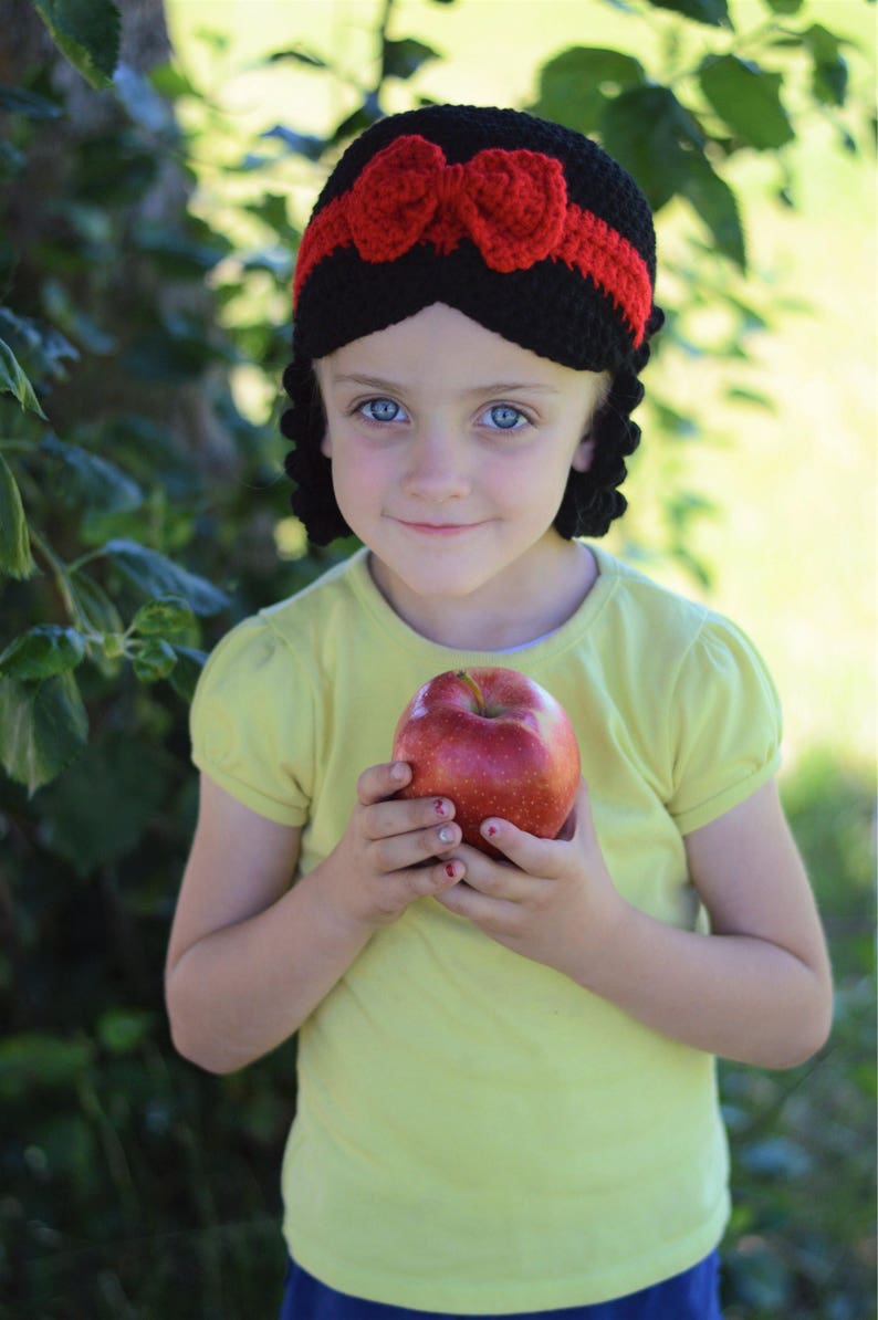 Puede incluir: Una diadema negra de ganchillo con lazo y ribete rojos, combinada con una camiseta amarilla y una manzana roja. La diadema tiene un dise&ntilde;o &uacute;nico, y la manzana se sostiene en las manos.