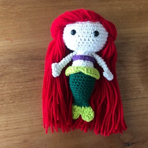 Pu&ograve; includere: Bambola sirena all'uncinetto con lunghi capelli rossi, un corpetto bianco, una parte superiore viola e verde e una coda verde.