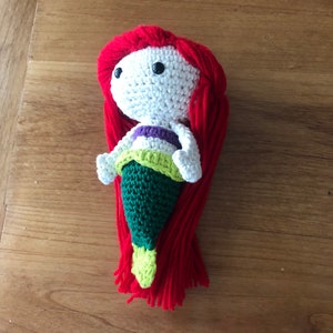 Pu&ograve; includere: Una bambola di sirena all'uncinetto con capelli di lana rossa, un corpo bianco, una parte superiore viola e verde e una coda verde.