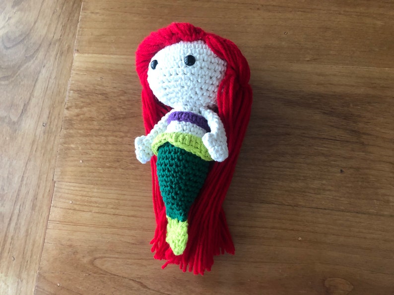 Pu&ograve; includere: Una bambola di sirena all'uncinetto con capelli di lana rossa, un corpo bianco, una parte superiore viola e verde e una coda verde.