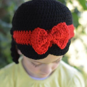 Puede incluir: Un gorro negro de ganchillo con una diadema roja y un lazo rojo. El gorro est&aacute; dise&ntilde;ado para parecerse a un personaje de cuento de hadas. El gorro lo lleva un ni&ntilde;o.