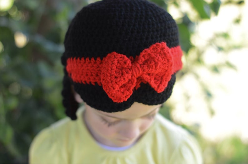 Puede incluir: Un gorro negro de ganchillo con una diadema roja y un lazo rojo. El gorro est&aacute; dise&ntilde;ado para parecerse a un personaje de cuento de hadas. El gorro lo lleva un ni&ntilde;o.