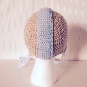 May include: A crocheted baby hat with a tan and light blue color scheme. The hat has two long ties that hang down the sides.