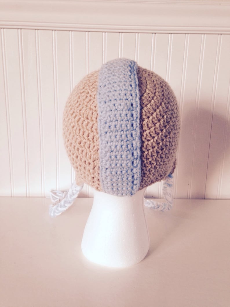 May include: A crocheted baby hat with a tan and light blue color scheme. The hat has two long ties that hang down the sides.