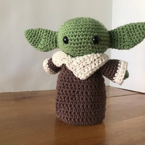May include: A crocheted green and brown plush toy with large ears and black eyes. The toy is wearing a white crocheted collar and brown crocheted sleeves.