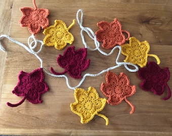 Pode incluir: Uma guirlanda de folhas de outono de crochê em tons de vermelho, laranja e amarelo. As folhas são conectadas por um cordão branco.