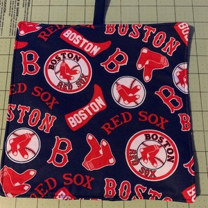 May include: A blue fabric pot holder with a red, white, and blue Boston Red Sox logo pattern. The pot holder has a loop handle.