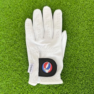 May include: White golf glove with a black and red emblem featuring a skull and lightning bolt design. The glove has small perforations for breathability and is set against a green background.