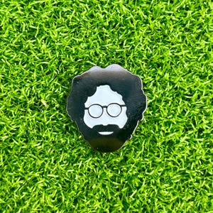 May include: A black and white pin featuring a stylized face with glasses, a beard, and curly hair. The pin is set against a background of bright green artificial turf.