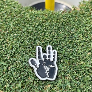 May include: A black and white golf ball marker in the shape of a hand with a unique design, resting on a green golf course. The marker is near the hole and a yellow flagstick.