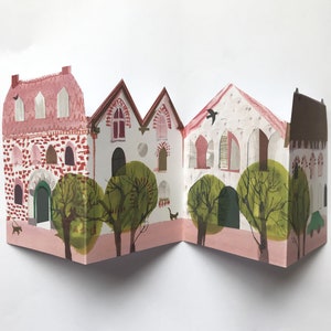 A concertina folded, shaped illustrated greetings card shpws a row of littel houses in pink, white and brown. Green trees line the street. The card lies against a white background.