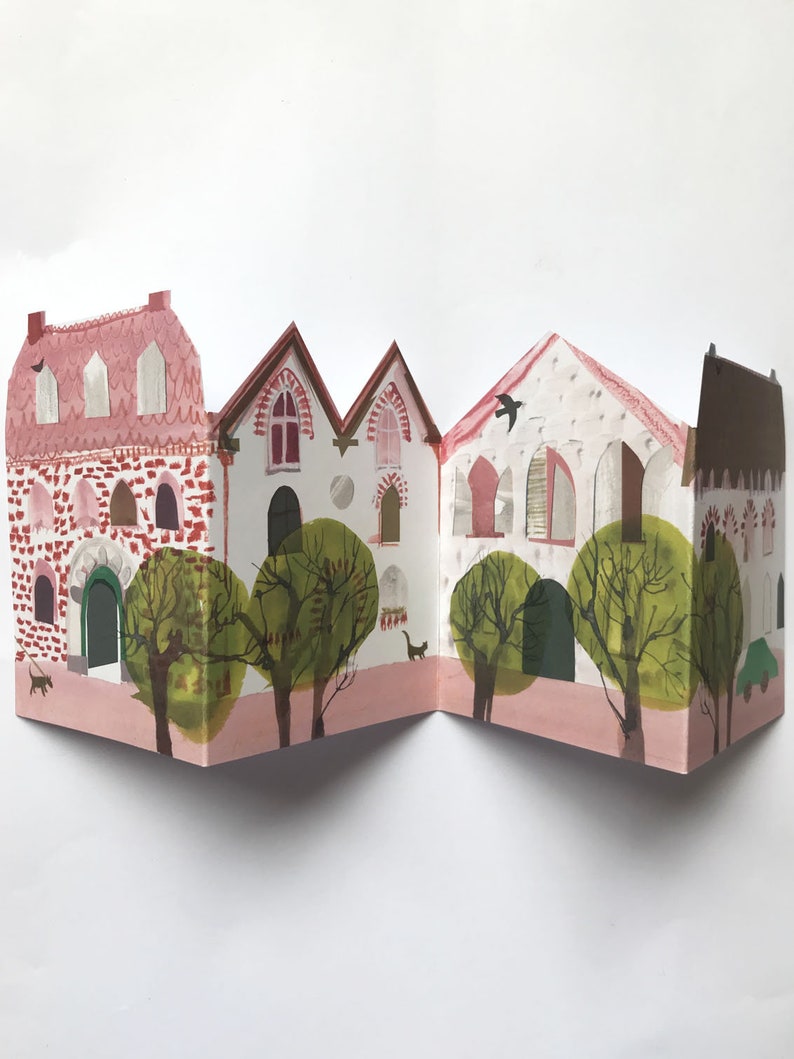 A concertina folded, shaped illustrated greetings card shpws a row of littel houses in pink, white and brown. Green trees line the street. The card lies against a white background.