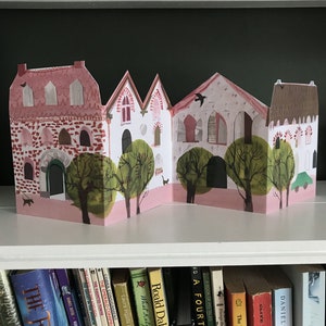 A concertina folded, shaped illustrated greetings card shpws a row of littel houses in pink, white and brown. Green trees line the street. The card sits on a shelf, above a row of books.
