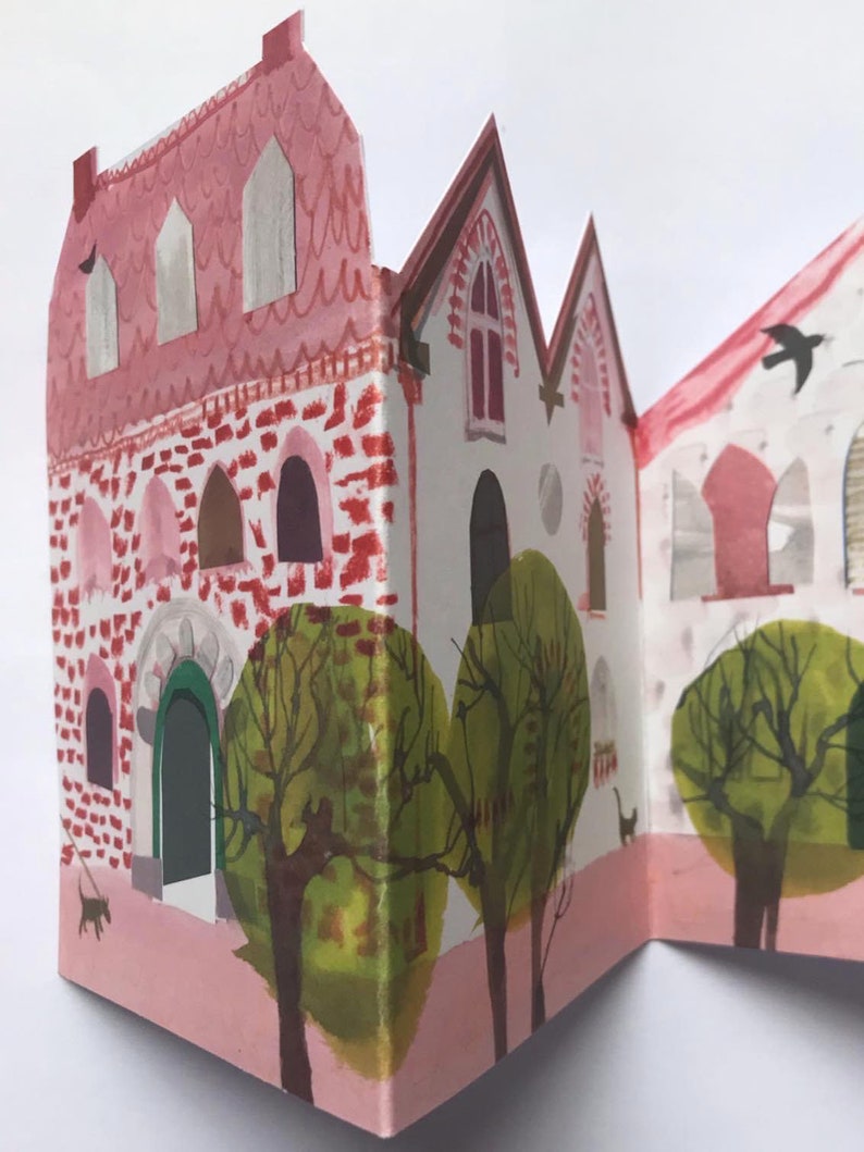 A concertina folded, shaped illustrated greetings card shpws a row of littel houses in pink, white and brown. Green trees line the street.