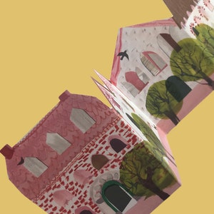 A concertina folded, shaped illustrated greetings card shpws a row of littel houses in pink, white and brown. Green trees line the street. The card is held diagonally by ajust-seen hand, against a yellow background.