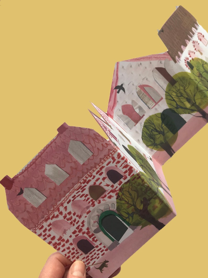 A concertina folded, shaped illustrated greetings card shpws a row of littel houses in pink, white and brown. Green trees line the street. The card is held diagonally by ajust-seen hand, against a yellow background.