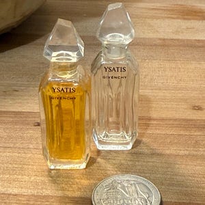 May include: Two miniature glass perfume bottles labeled Ysatis Givenchy, one filled with amber liquid and one empty, featuring faceted clear stoppers and hexagonal bodies, positioned next to a United States quarter coin for scale.