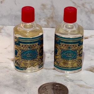 Pode incluir: Duas pequenas garrafas de vidro transparente de água de colónia 4711 com tampas de rosca vermelho-brilhante. Cada garrafa apresenta um rótulo decorativo dourado e azul-petróleo com o texto 4711 e Glockengasse No 4711. Uma moeda está à frente.