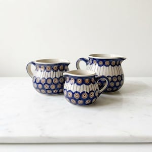 May include: Three ceramic creamers featuring a blue and brown circular pattern on a white background. Each piece has a rounded body, a small pouring spout, and a curved handle. The design consists of repeating concentric circles.