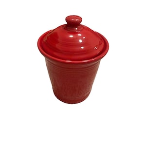 Puede incluir: Un recipiente de cerámica roja con tapa. El recipiente tiene forma cilíndrica con un borde ligeramente ensanchado y una serie de estrías horizontales. La tapa es redondeada con un pequeño pomo en la parte superior. El recipiente es de color rojo brillante.