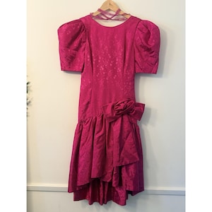 May include: A bright pink vintage dress with short, puffed sleeves and a large bow at the waist. The dress has a high-low hemline and a floral pattern. It is hanging on a wooden hanger.