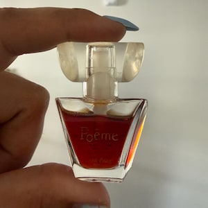 Puede incluir: Un pequeño frasco de perfume de vidrio transparente, con una tapa decorativa. El frasco contiene un líquido marrón rojizo y presenta la palabra "Poême". El frasco se sostiene entre dos dedos.