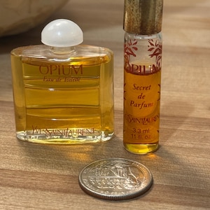 May include: Two glass bottles of Yves Saint Laurent Opium fragrance with amber liquid. One is a wide, flat bottle labeled Eau de Toilette, and the other is a tall, thin vial labeled Secret de Parfum 3.3 ml, 0.11 fl. oz. A US quarter coin is placed in front.