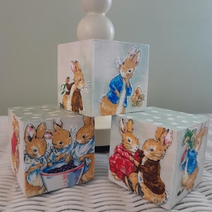 May include: Three wooden blocks with illustrations of Peter Rabbit characters. The blocks are painted with a light blue background and white polka dots. The illustrations feature rabbits in various outfits, holding a bowl of blueberries and carrying vegetables.