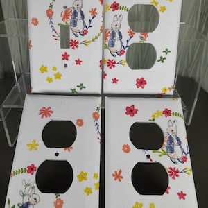 May include: Four white light switch and outlet covers with a floral pattern featuring a bunny rabbit. The bunny is wearing a blue jacket and has a white tail. The flowers are in shades of pink, yellow, and green.