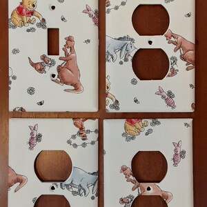 May include: Four white electrical outlet and switch covers featuring illustrations of Winnie the Pooh and friends. The covers have a repeating pattern of characters and floral accents, with brown openings for outlets and switches.