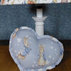 May include: A heart-shaped pillow featuring illustrations of Peter Rabbit on a light blue background with white stars. The pillow has a scalloped edge and is placed on a wooden surface. A matching lampshade with rabbit illustrations is visible.