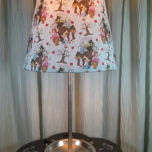 May include: A silver table lamp with a white lampshade featuring a whimsical pattern of characters from the Wizard of Oz. The lampshade has a blue background with red poppy flowers and green leaves.