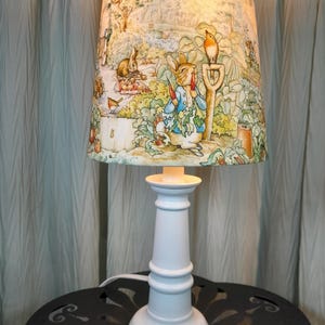 May include: A white table lamp with a lampshade featuring a colorful illustration of rabbits and a garden scene. The lamp has a white base and a decorative lampshade with a detailed storybook design.