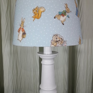 May include: A white table lamp with a blue and white patterned lampshade featuring Peter Rabbit and other characters. The lampshade has white polka dots on a light blue background.