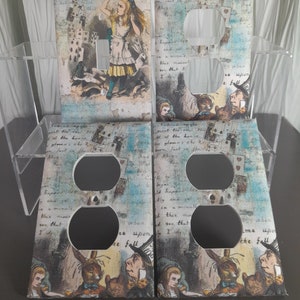 May include: Four decorative light switch and outlet covers featuring a vintage Alice in Wonderland illustration with a blue, white, and brown color scheme. The text "I tried then all" is visible on the top left cover. The text "I have to see that" is visible on the top right cover. The text "where upon the fall" is visible on the bottom left cover. The text "where upon the fall" is visible on the bottom right cover.