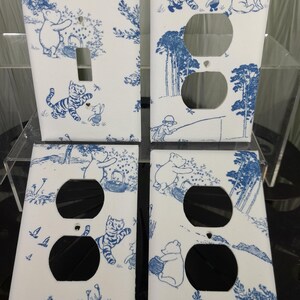 May include: Set of four Winnie the Pooh themed light switch and outlet covers. The covers feature a white background with blue illustrations of Winnie the Pooh, Tigger, Piglet, and Eeyore in a woodland setting.