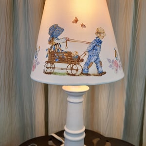 May include: White ceramic table lamp with a white fabric lampshade featuring a printed design of two children pulling a wagon with butterflies.