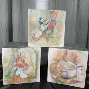 May include: Three square wooden blocks with illustrations from a children's story. The blocks feature images of a fox, a duck, rabbits, and a rabbit in a red cape. The blocks are green with white polka dots on the top.