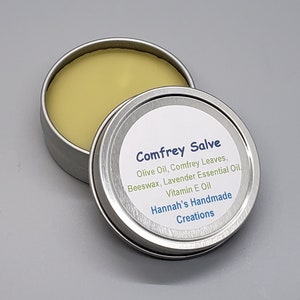 May include: A silver tin with a white label that reads "Comfrey Salve Olive Oil, Comfrey Leaves, Beeswax, Lavender Essential Oil, Vitamin E Oil Hannah's Handmade Creations". The tin is open and contains a yellow salve.