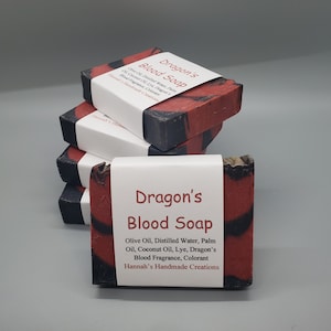 May include: A stack of four bars of Dragon's Blood Soap. The soap is red and black with a label that reads "Dragon's Blood Soap" and lists the ingredients: Olive Oil, Distilled Water, Palm Oil, Coconut Oil, Lye, Dragon's Blood Fragrance, Colorant. The label also includes the text "Hannah's Handmade Creations".