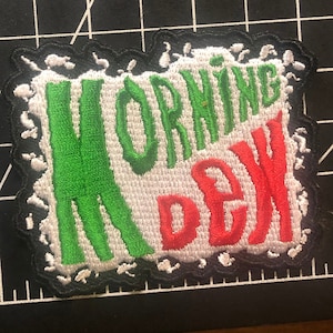 May include: A white, green, and red embroidered patch with the words "MORNING DEW" in a stylized font. The patch has a black, jagged border.